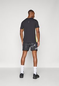 Schwarz sportliches T-Shirt mit gelben seitlichen Akzenten, kombiniert mit grauen gemusterten Shorts. Weiße Socken und schwarze Sneaker runden den Look ab.