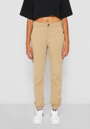 Woman wearing beige cargo pants with elastic cuffs, white Nike sneakers, and a black cropped top, standing against a plain light gray background.