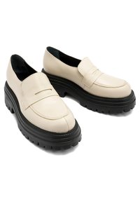 Beżowe skórzane mokasyny z grubą czarną gumową podeszwą, z wyszywaną końcówką oraz detalem w postaci paska z przodu.