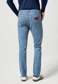 Jeans de mezclilla azul claro con piernas estrechas, bolsillos traseros con un diseño bordado y una etiqueta de marca de piel marrón en la cintura.
