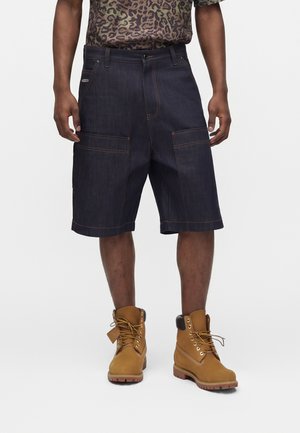 Muž nosi tmavé denim cargo šortky, svetlohnedé šnurovacie pracovné topánky a košeľu s leopardím vzorom, stojí pred jednoduchým pozadím.