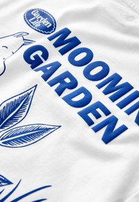 Vit tygtröja med blå text som säger "MOOMIN GARDEN" och stiliserade blå blad, med en rund logotyp ovanför. Mjuk textur och avslappnad design.