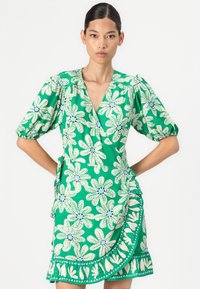 Groene wikkeljurk met korte puffmouwen, voorzien van witte bloemenpatronen en blauwe accenten. De gerimpelde zoom voegt textuur en detail toe.