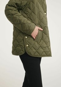 Oliwkowa pikowana kurtka w romby, z boczną kieszenią i zapięciem na napy. Miękka tekstura i kontrastowe wykończenie u dołu.