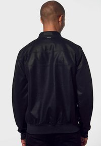 Giacca bomber nera con finitura liscia, polsini e orlo a coste, e un piccolo etichetta sul retro. Design semplice e classico con una vestibilità su misura.