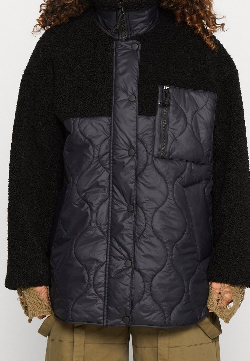 Veste matelassée noire avec boutons-pression et poche poitrine zippée, associée à des manches et un col en polaire noirs, portée avec un pantalon cargo beige.