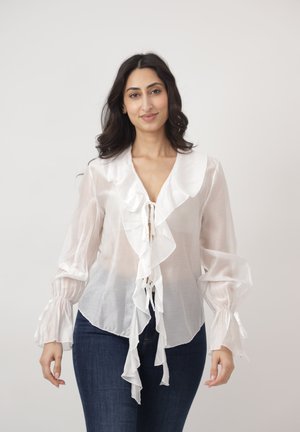 Kobieta o długich ciemnych włosach, ubrana w przezroczystą białą bluzkę z falbanami i ciemnoniebieskie dżinsy, stojąca na tle jednolitego, jasnego tła.