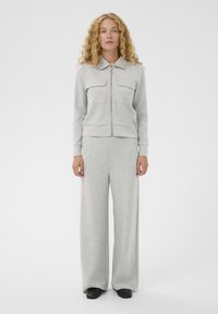 Žena s kučeravými vlasmi, oblečená vo svetlosivej mikine na zips a širokých nohaviciach, stojí proti jednoduchému bielému pozadiu.