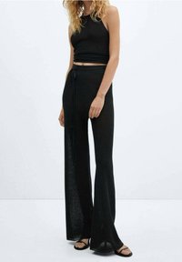 Brassière noire côtelée associée à un pantalon noir ample et évasé, doté d'une taille à lien et d'une texture transparente, porté avec des sandales plates.