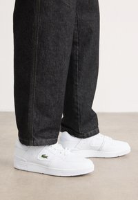 Bijele tenisice od kože, s perforiranim prstom, minimalističkim dizajnom i Lacoste logotipom. Nosi se s tamnim trapericama.