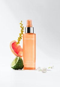 Pomarańczowa butelka Clinique Happy body spritz z srebrną nakrętką, otoczona różowym grejpfrutem, zieloną limonką i żółtymi kwiatowymi akcentami.