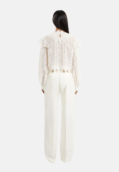 Kanten blouse met rimpelachtige schouders, roomkleurig, en een keyhole detail op de rug, gecombineerd met op maat gemaakte witte hooggetailleerde broek.