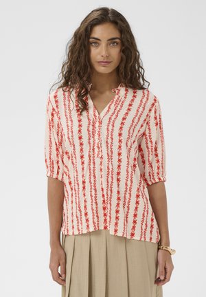 Barna göndör hajú nő fehér blúzban, amelyen piros, függőleges virágmintás csíkok vannak, és bézs színű, pliszírozott szoknyában, egyszínű háttér előtt áll.