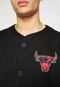 Camisa de botones negra a rayas con un logo gráfico de un toro rojo y blanco en el pecho. La tela tiene una textura suave.