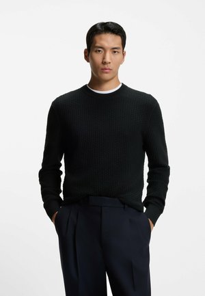 Maglione nero in maglia a trama con scollo rotondo, maniche lunghe e polsini a coste, abbinato a pantaloni scuri sartoriali.