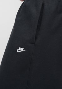 Μαύρο παντελόνι φόρμας Nike με λευκό λογότυπο στη αριστερή πλευρά, κατασκευασμένο από απαλό ύφασμα με άνετη εφαρμογή και λεπτομέρεια πλαϊνής τσέπης.