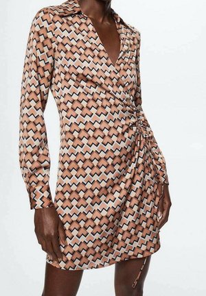 Kvinde iført en minikjole med lange ærmer i en geometrisk mønster med farverne tan, sort, hvid og lyseblå samt med en rynkedetalje på højre side.