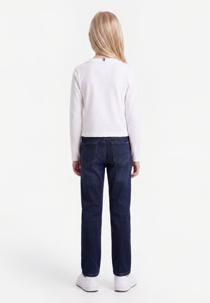 Enfant aux longs cheveux blonds portant une chemise blanche à manches longues, un jean bleu foncé et des baskets blanches, debout de dos.