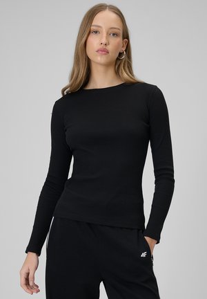 Young woman with long hair wears a fitted black long-sleeve top and black pants, standing against a plain light gray background.
