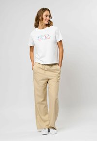 Witte katoenen t-shirt met kleurrijke florale graphics op de voorkant, gecombineerd met beige wijde broek en witte sneakers.