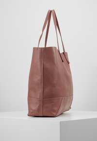 Vaaleanpunainen nahkainen olkalaukku, jossa on kaksi pitkää kahvaa, jäsennelty muoto ja näkyvät tikkaus-yksityiskohdat saumoissa ja pohjassa.