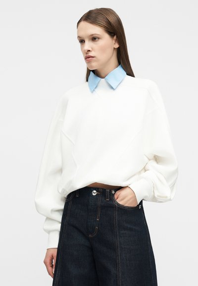 Γυναίκα με ίσια καστανά μαλλιά φοράει λευκό φαρδύ φούτερ με μπλε γιακά και σκούρο τζιν ψηλόμεσο με φαρδιά μπατζάκια, χέρι στην τσέπη.