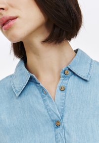 Jasnoniebieska koszula chambray z kołnierzykiem, z dwoma drewnianymi guzikami i lekko teksturowanym materiałem. Zbliżenie.