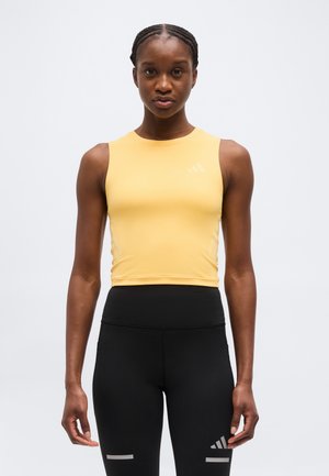 Športovo založená žena s vrkočmi má na sebe žltý crop top bez rukávov a čierne legíny s vysokým pásom a reflexnými logami.