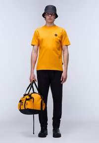 Camiseta amarilla de manga corta con logo, sombrero tipo cubo negro, pantalones negros y una bolsa de deporte amarilla y negra con asas. Texturas suaves en todo.