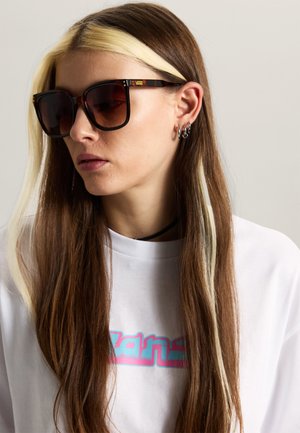 Jong persoon met lang bruin haar en blonde strepen draagt grote bruine Vans-zonnebril en wit shirt met kleurrijk logo.