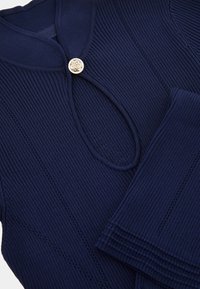 Mörkblå ribbad stickad topp med rund hals och nyckelhålsdetalj, försedd med en dekorativ silverknapp och texturerade ärmslut.