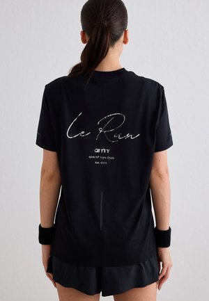 Kobieta z ciemnym kucykiem, ubrana w czarną koszulkę "Le Run" i opaski na nadgarstkach, zwrócona tyłem, na jednolitym jasnym tle.