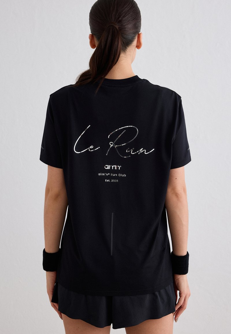 Femme avec queue de cheval foncée portant un t-shirt noir "Le Run" et des bracelets, vue de dos, sur fond clair uni.