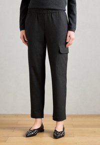 Un pantalon gris foncé à rayures avec une taille élastique et une poche latérale. Porté avec des ballerines noires cloutées sur un sol en bois.