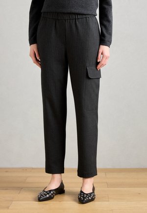Pantaloni a righe grigio scuro con elastico in vita e tasca laterale. Indossati con ballerine nere con borchie su un pavimento di legno.