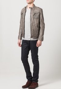 Giacca in pelle grigia da uomo con due tasche sul petto, chiusura con zip e dettagli testurizzati, abbinata a jeans scuri e scarpe marroni.