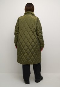 Caban matlasat de culoare verde măsliniu, cu guler înalt, având o siluetă lungă și un model în diamante, realizat dintr-o țesătură lucioasă, asortat cu pantaloni negri.