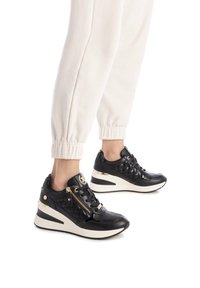 Zapatillas altas negras con textura acolchada, acentos dorados, cremalleras laterales y suela de cuña, combinadas con joggers color crema.