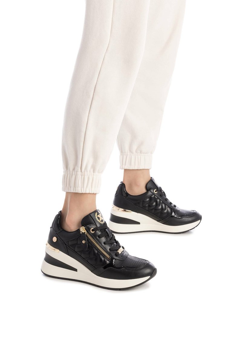 Zapatillas altas negras con textura acolchada, acentos dorados, cremalleras laterales y suela de cuña, combinadas con joggers color crema.