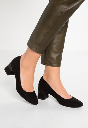 Chaussures à talons blocs en daim noir avec un bout arrondi, présentant un motif texturé à l'arrière du talon. Associées à un pantalon vert olive.