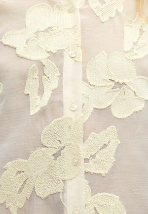 Blouse à boutons pour femmes ornée d'une délicate broderie florale blanche sur un fond beige clair, avec une texture délicate et des boutons nacrés.