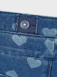 Tela de denim azul con un patrón de corazones. Detalle que incluye un botón de plástico transparente y costuras en contraste en la cinturilla.