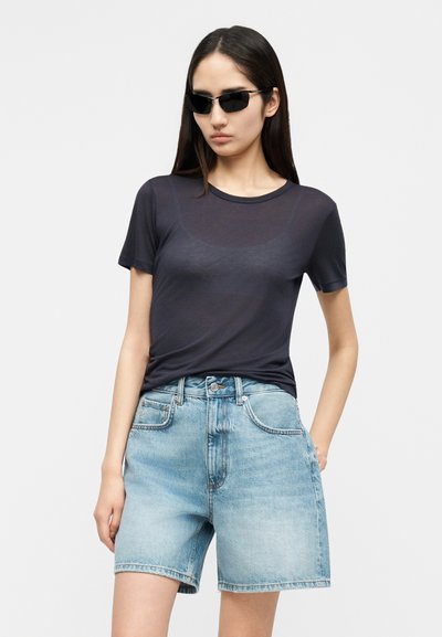 Vrouw met zwarte zonnebril, doorschijnend zwart shirt met korte mouwen en lichtblauwe denim shorts met hoge taille, staand tegen een effen achtergrond.