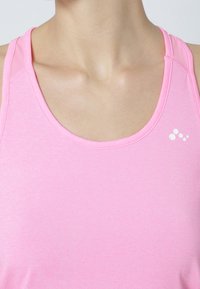 Różowy sportowy top na ramiączkach wykonany z gładkiej tkaniny, z dekoltem w serek i małym białym logo po lewej stronie.