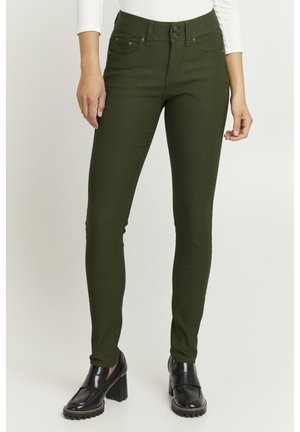 Schmale grüne Jeans mit glatter Textur, fünf Taschen und Doppelknopfverschluss. Getragen mit schwarzen Plateauschuhen.
