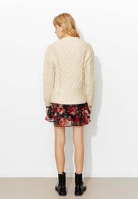 Pull crème à torsades avec un motif en losange texturé, porté sur une robe noire à fleurs avec volants. Des bottines noires complètent la tenue.