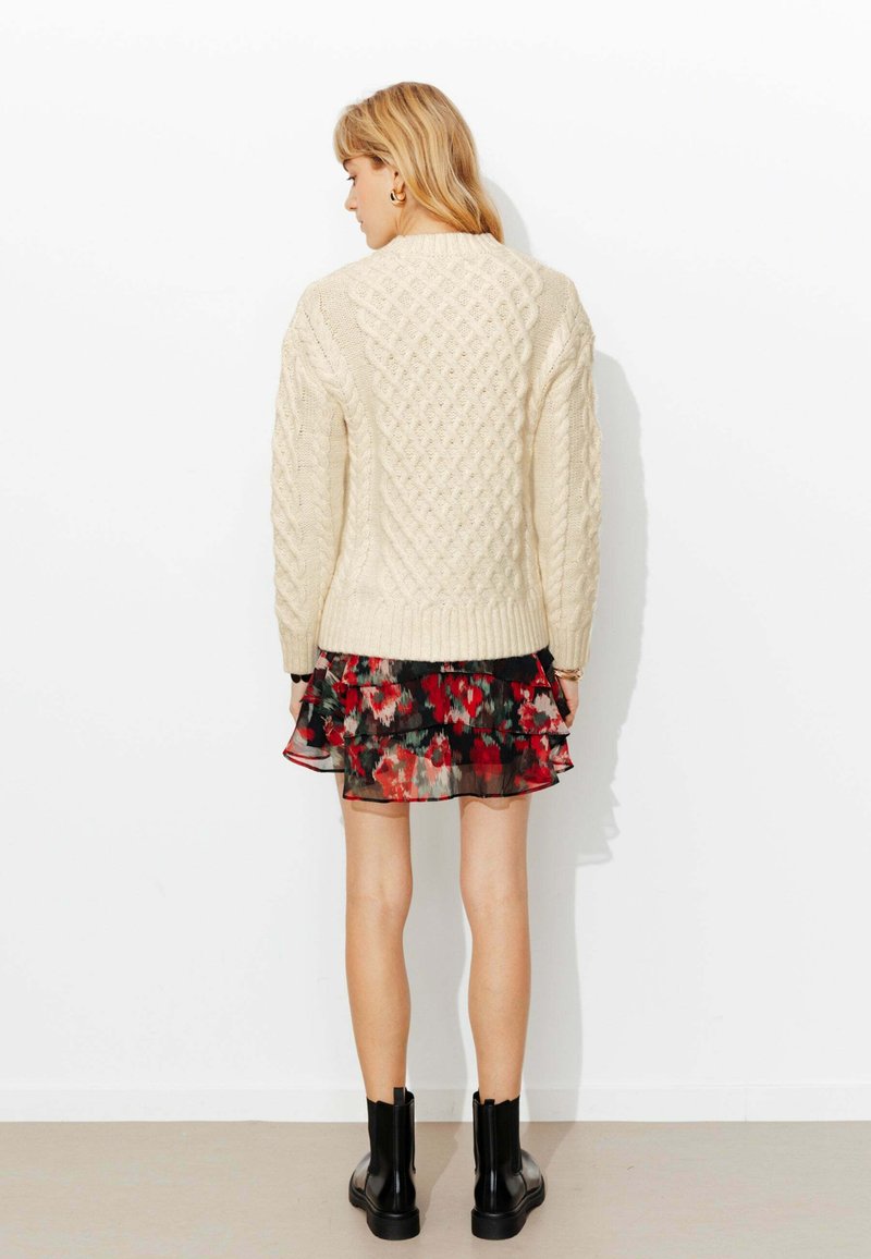 Pull crème à torsades avec un motif en losange texturé, porté sur une robe noire à fleurs avec volants. Des bottines noires complètent la tenue.