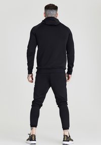 Felpa nera con design aderente, dotata di tasca a marsupio e maniche raglan. Abbinata a pantaloni sportivi coordinati e sneakers nere.