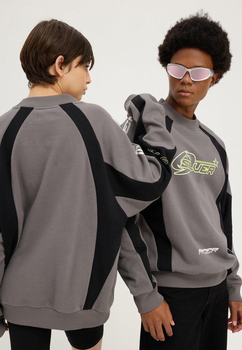 Deux personnes portant des sweat-shirts assortis gris et noirs, l'une de profil et l'autre portant des lunettes de soleil réfléchissantes roses, posant devant un fond uni.