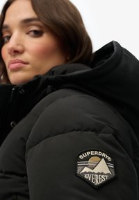 Sort dunjakke med høj krave og et logo-patch på venstre skulder med bjerge og teksten "SUPERDRY EVEREST".